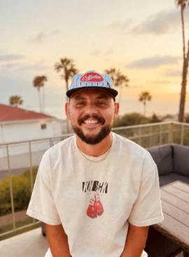 Cory sitting on a deck at sunset with palm trees and the coast behind him.
