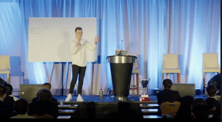 Cory presenting on a lit stage beside a whiteboard, teaching to a seated audience.